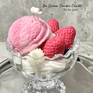 May include: A pink and white ice cream sundae candle in a clear glass dish. The candle is shaped like a sundae with a scoop of ice cream, whipped cream, and strawberries. The candle is labeled "Ice Cream Sundae Candle C&L Art Space".
