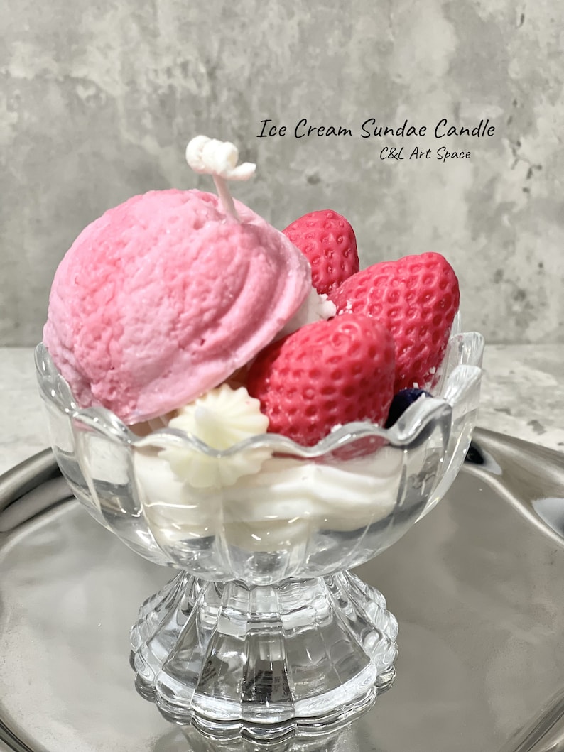 May include: A pink and white ice cream sundae candle in a clear glass dish. The candle is shaped like a sundae with a scoop of ice cream, whipped cream, and strawberries. The candle is labeled "Ice Cream Sundae Candle C&L Art Space".