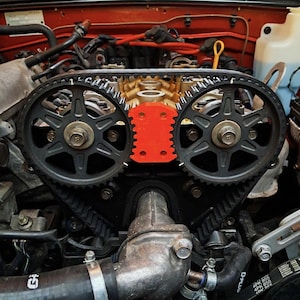 May include: Detailed view of a car engine's internal workings. Two large, dark grey gears are linked by a black belt. A bright orange component is positioned between the gears. The background shows red and metallic engine parts.