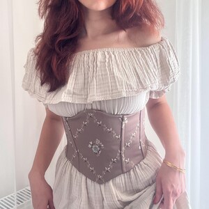 K&ouml;nnte beinhalten: Beiges schulterfreies Oberteil und Kleid mit braunem Korsett. Das Korsett hat Blumenstickereien und ein Rautenmuster. Ein goldenes Armband ist am Handgelenk zu sehen.