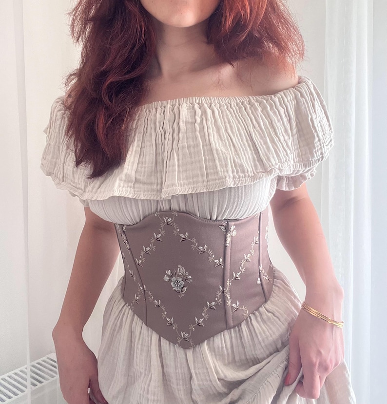 K&ouml;nnte beinhalten: Beiges schulterfreies Oberteil und Kleid mit braunem Korsett. Das Korsett hat Blumenstickereien und ein Rautenmuster. Ein goldenes Armband ist am Handgelenk zu sehen.
