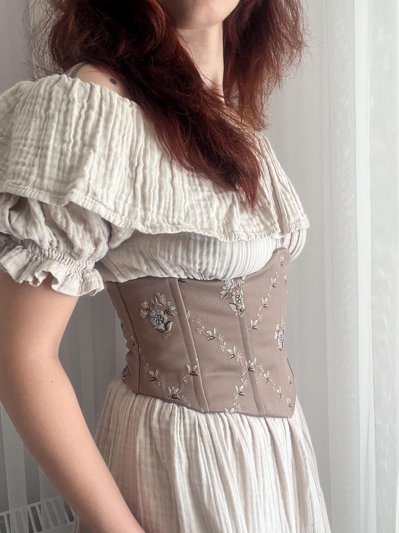 K&ouml;nnte beinhalten: Beiges Oberteil und Kleid mit freier Schulter. &Uuml;ber dem Kleid wird ein braunes Korsett mit floraler Stickerei getragen. Das Outfit besteht aus einem weichen, strukturierten Stoff, der eine romantische, Vintage-&Auml;sthetik erzeugt. Der Gesamtstil erinnert an historische Mode.