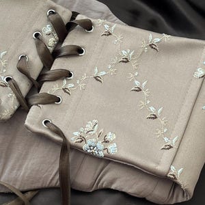 K&ouml;nnte beinhalten: Ein beiges Korsett mit floraler Stickerei und brauner Bandschn&uuml;rung. Das Korsett hat detaillierte gestickte Blumenmuster in Wei&szlig;, Braun und Blau. Das braune Band ist durch Metall&ouml;sen gef&auml;delt.