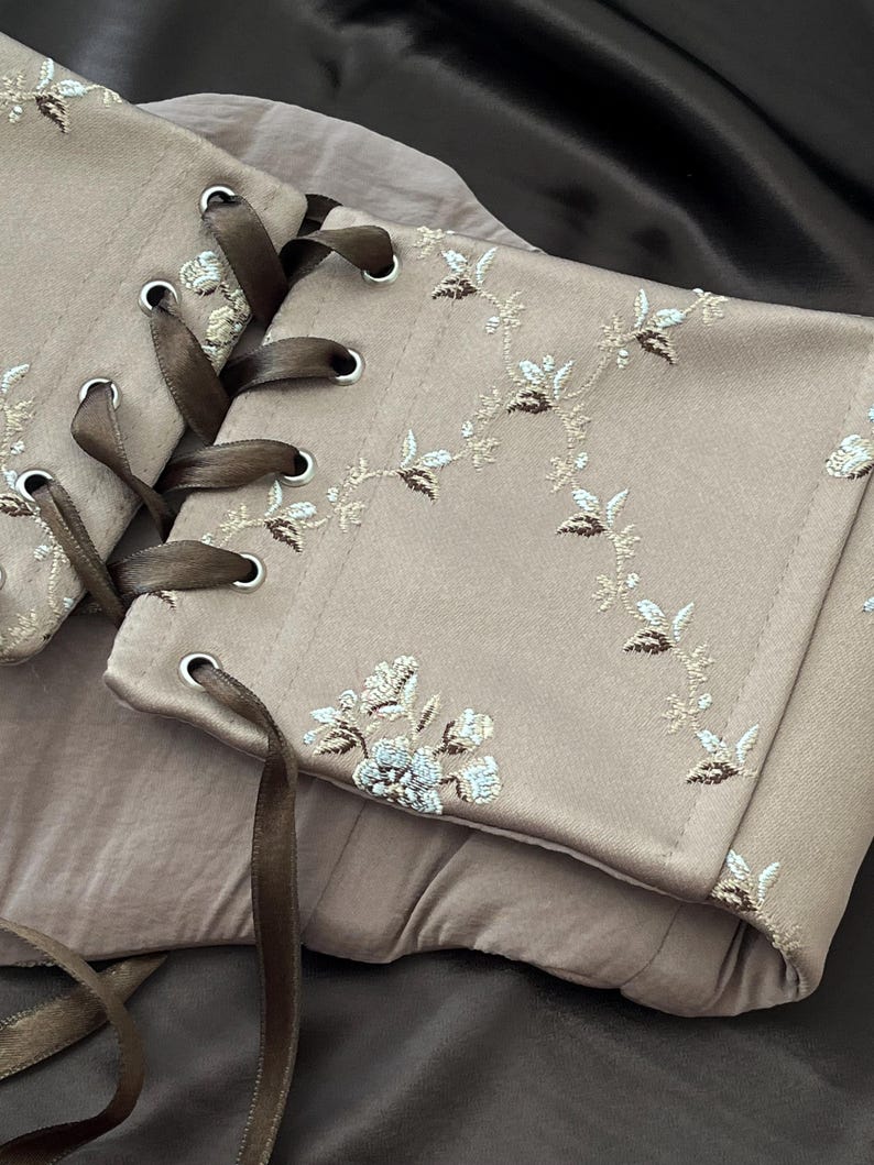 K&ouml;nnte beinhalten: Ein beiges Korsett mit floraler Stickerei und brauner Bandschn&uuml;rung. Das Korsett hat detaillierte gestickte Blumenmuster in Wei&szlig;, Braun und Blau. Das braune Band ist durch Metall&ouml;sen gef&auml;delt.