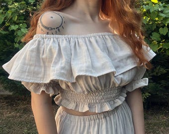 Könnte beinhalten: Ein beiges schulterfreies Crop-Top mit Rüschenärmeln und einem passenden hoch taillierten Rock. Das Oberteil hat einen gerafften Bund und ein mehrschichtiges Rüschendesign. Das Outfit ist aus leichtem Stoff gefertigt, perfekt für warmes Wetter.