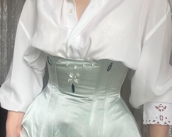 Könnte beinhalten: Ein hellgrünes Satin-Korsett mit dekorativen Verzierungen. Das Korsett hat ein strukturiertes Design mit vertikalen Nähten und einer leichten Kurve an der Taille. Es wird mit einem weißen Hemd mit Knöpfen getragen.