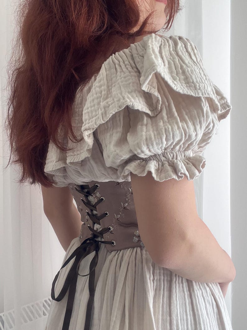 K&ouml;nnte beinhalten: Beiges, mehrschichtiges Kleid mit Korsett. Das Korsett ist braun mit floraler Stickerei und schwarzer Schn&uuml;rung. Das Kleid hat einen ger&uuml;schten Ausschnitt und &Auml;rmel. Der Stoff scheint ein weiches, strukturiertes Material zu sein.