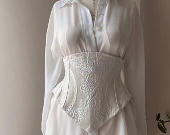 May include: A white long-sleeved button-down shirt with a pointed collar featuring lace trim, worn under a structured white corset with a raised, intricate tonal damask pattern and a pointed hemline that sits at the waist.