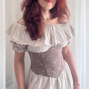 K&ouml;nnte beinhalten: Beiges schulterfreies Kleid mit ger&uuml;schtem Ausschnitt und &Auml;rmeln. Das Kleid hat ein tailliertes, taupefarbenes Korsett mit gestickten Blumenmustern, das eine romantische und Vintage-&Auml;sthetik erzeugt.