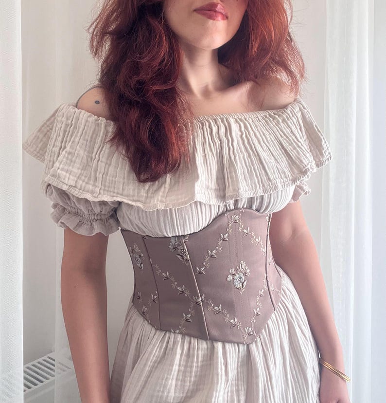 K&ouml;nnte beinhalten: Beiges schulterfreies Kleid mit ger&uuml;schtem Ausschnitt und &Auml;rmeln. Das Kleid hat ein tailliertes, taupefarbenes Korsett mit gestickten Blumenmustern, das eine romantische und Vintage-&Auml;sthetik erzeugt.