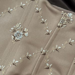 K&ouml;nnte beinhalten: Nahaufnahme eines beigefarbenen Korsetts mit floraler Stickerei. Die Stickerei zeigt wei&szlig;e, hellbraune und goldfarbene Blumen und Bl&auml;tter. Das Korsett hat vertikale N&auml;hte und eine geschwungene Form, was auf ein eng anliegendes Kleidungsst&uuml;ck hindeutet.
