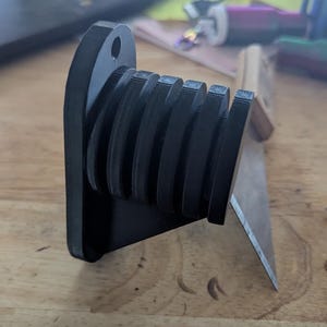 May include: A black, 3D-printed object with a series of curved, stacked elements. The object has a flat base with a hole and is positioned on a wooden surface. A metal blade is visible in the background.