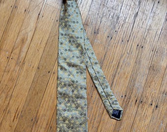 May include: A patterned tie in gold, olive green, and grey, knotted at the top. The tie has a repeating floral design and diagonal stripes. The tie is lying on a wooden surface.
