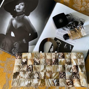 May include: A mosaic-patterned clutch bag in beige, brown, and cream tones, with a curved handle. It's displayed on a magazine featuring a black and white photograph of a woman wearing a hat and other accessories.