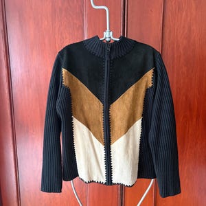 May include: A black, brown, and cream suede jacket with a ribbed knit collar and sleeves. The jacket has a chevron design on the front and a zip closure. It is hanging on a metal hanger.
