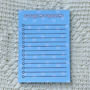 Puede incluir: Un bloc de notas azul claro con el texto "FUTURE ME PROBLEMS" en negro. El bloc de notas tiene líneas horizontales para escribir e ilustraciones de pequeñas flores. El bloc de notas está sobre una superficie blanca texturizada.