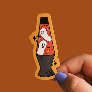 May include: A sticker of a lava lamp with a Halloween theme. The lamp is orange and red with a black base and two white ghosts inside. The sticker is on a brown background.