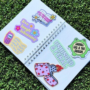 May include: A white notebook with colourful stickers on it. The stickers feature phrases like "Live laugh lobotomy", "Skip the flowers buy the book", "Unhinged & Feral", and "Sounds like a you problem".
