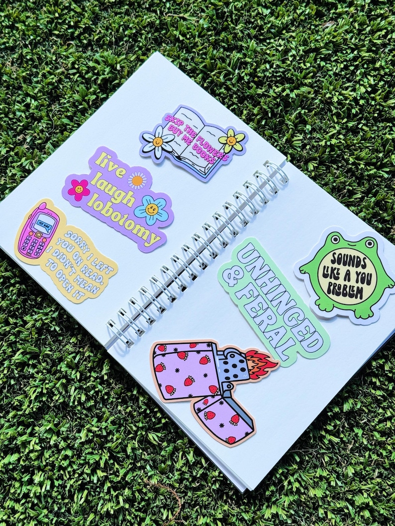 May include: A white notebook with colourful stickers on it. The stickers feature phrases like "Live laugh lobotomy", "Skip the flowers buy the book", "Unhinged & Feral", and "Sounds like a you problem".