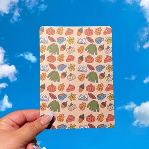 Könnte beinhalten: Eine rechteckige Karte mit beigem Hintergrund und einem sich wiederholenden Muster von Herbstillustrationen. Das Muster umfasst Kürbisse, Pullover, Kaffeetassen, Kuchenstücke, Eicheln und Herbstblätter. Die Karte wird vor einem blauen Himmel gehalten.
