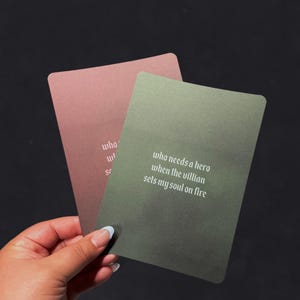 Könnte beinhalten: Zwei rechteckige Karten, eine staubrosa und eine olivgrüne, vor dunklem Hintergrund. Auf der grünen Karte steht "who needs a hero when the villain sets my soul on fire." Die staubrosa Karte hat teilweise Text.