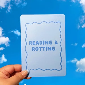 May include: A light blue card with the words "READING & ROTTING" in a playful font, framed by a wavy border. The card is held against a backdrop of a bright blue sky with fluffy white clouds.