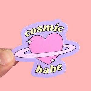 May include: Purple sticker with a pink heart surrounded by a white ring, with the text "cosmic babe" in yellow letters.