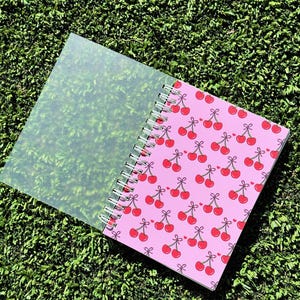 May include: A pink spiral bound notebook with a cherry pattern. The notebook has a clear plastic cover.