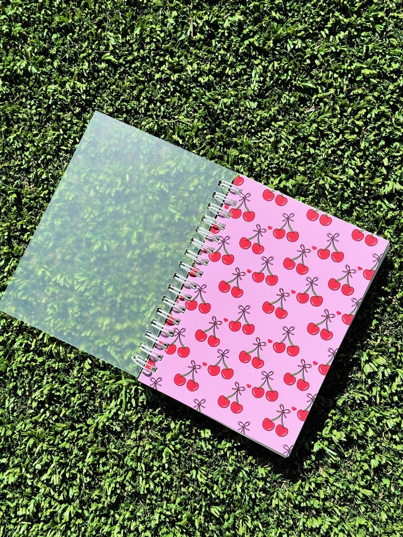 May include: A pink spiral bound notebook with a cherry pattern. The notebook has a clear plastic cover.