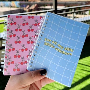 May include: Two small spiral notebooks. One has a pink cover with a repeating pattern of red cherries. The other has a blue cover with a grid pattern and the text "For all my silly little stickers".