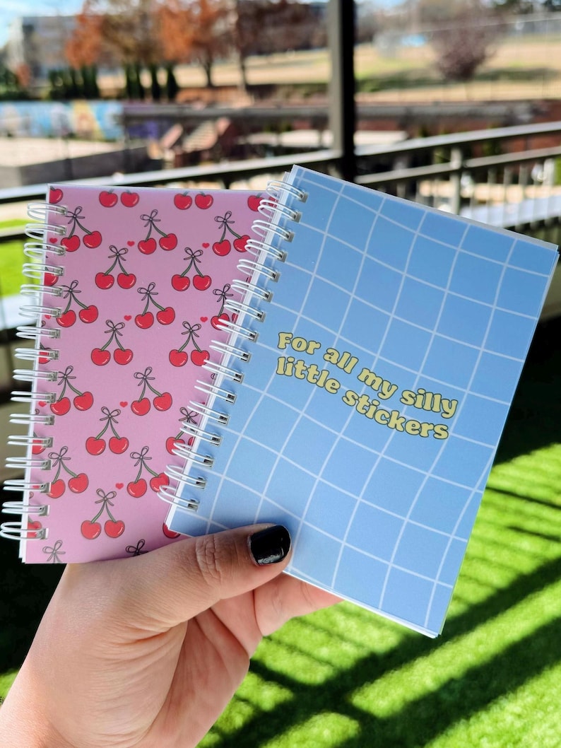 May include: Two small spiral notebooks. One has a pink cover with a repeating pattern of red cherries. The other has a blue cover with a grid pattern and the text "For all my silly little stickers".