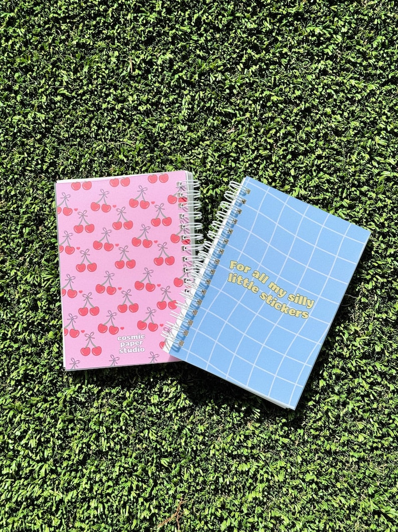 May include: Two spiral bound notebooks. One is pink with a cherry pattern and the text "cosmic paper studio". The other is blue with a grid pattern and the text "For all my silly little stickers".