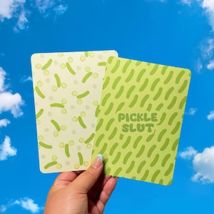 May include: Two rectangular cards with pickle-themed designs. One card features a pattern of pickles and pickle slices on a light yellow background. The other card has a green background with a pickle pattern and the text "PICKLE SLUT".