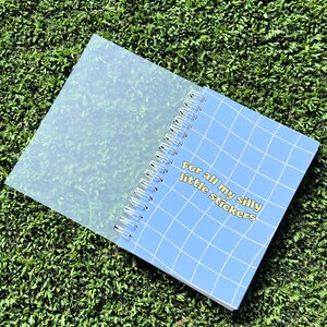 May include: A blue spiral bound notebook with a grid pattern and the text "For all my silly little stickers" in yellow. The notebook has a clear plastic cover.