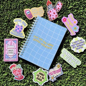 May include: A blue notebook with a grid pattern and the text "For all my silly little stickers" on the cover. The notebook is surrounded by colourful stickers with various designs and text, including "Crying over fictional characters", "Sorry I left you on read I didn't mean to open it", "Live laugh lobotomy", "I can't so out tonight my tummy hurts", "Unhinged & Feral", "Sounds like a you problem", "Disco Strawberry", "Stop the flowers but buy me books", and a lighter with a flame.