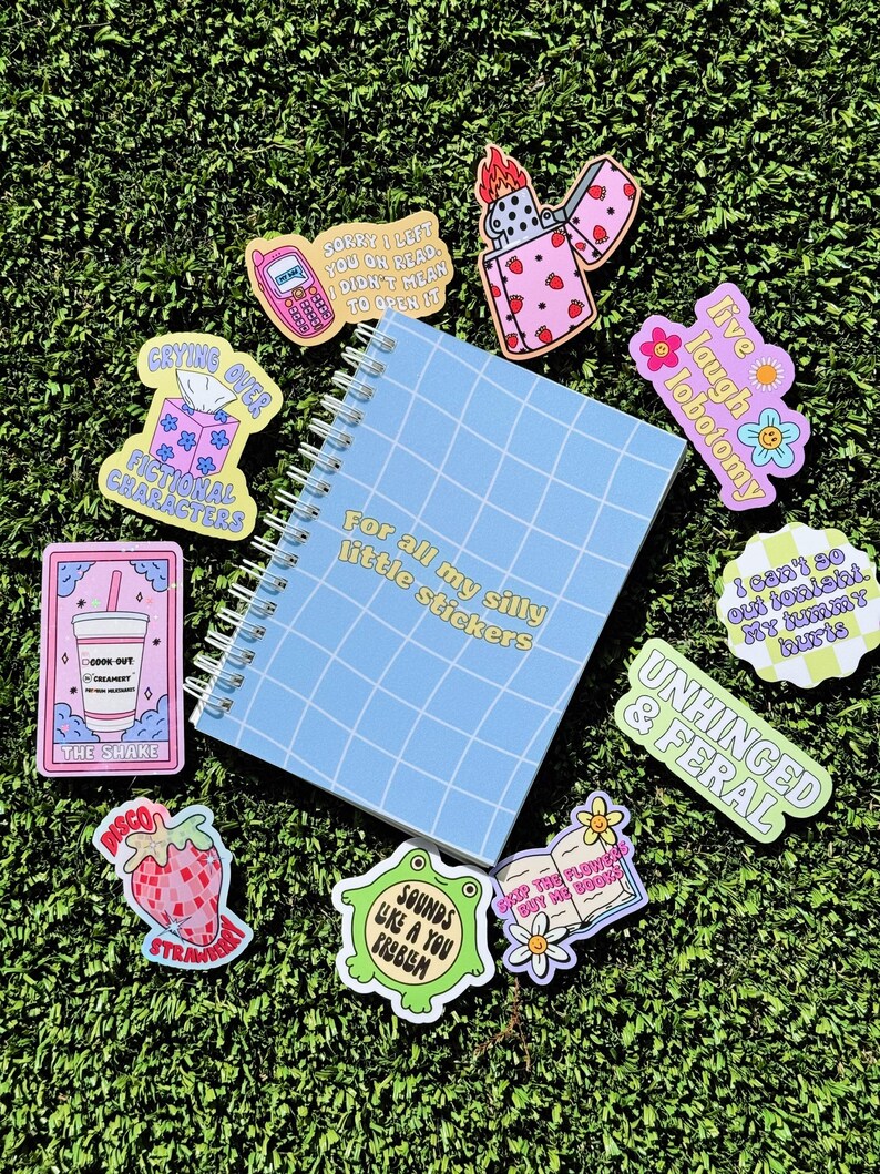 May include: A blue notebook with a grid pattern and the text "For all my silly little stickers" on the cover. The notebook is surrounded by colourful stickers with various designs and text, including "Crying over fictional characters", "Sorry I left you on read I didn't mean to open it", "Live laugh lobotomy", "I can't so out tonight my tummy hurts", "Unhinged & Feral", "Sounds like a you problem", "Disco Strawberry", "Stop the flowers but buy me books", and a lighter with a flame.
