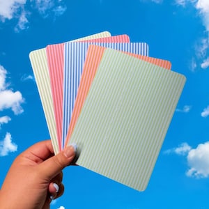 May include: A hand holding a fan of rectangular cards with vertical stripes in pastel colors: light green, pink, blue, and orange. The background is a bright blue sky with fluffy white clouds.