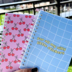 May include: Two small spiral notebooks. One has a pink cover with a repeating pattern of red cherries. The other has a blue cover with a grid pattern and the text "For all my silly little stickers".