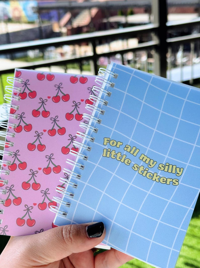 May include: Two small spiral notebooks. One has a pink cover with a repeating pattern of red cherries. The other has a blue cover with a grid pattern and the text "For all my silly little stickers".