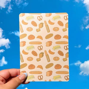 May include: A rectangular card featuring a repeating pattern of bread illustrations, including croissants, pretzels, and loaves, along with butter sticks. The card is held against a backdrop of a blue sky with fluffy white clouds.
