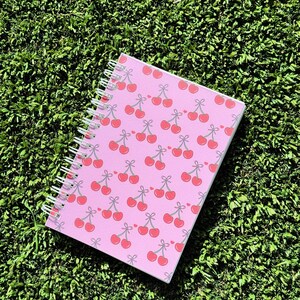 May include: A pink spiral notebook with a cherry pattern. The cherries are red with green stems and leaves.
