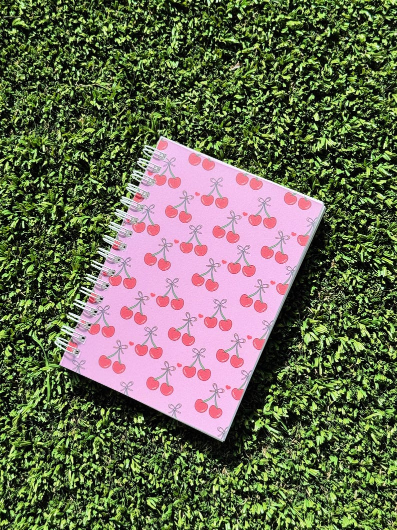 May include: A pink spiral notebook with a cherry pattern. The cherries are red with green stems and leaves.