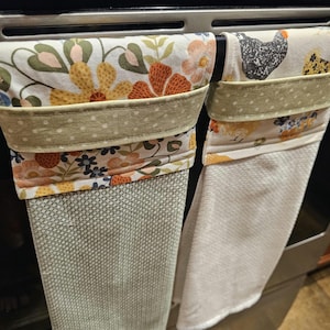 May include: Two hanging kitchen towels. One features a floral pattern with orange, yellow, and blue flowers. The other has a chicken print. Both have a green polka dot trim and are hanging on a metal bar.
