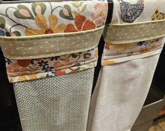 May include: Two hanging kitchen towels. One features a floral pattern with orange, yellow, and blue flowers. The other has a chicken print. Both have a green polka dot trim and are hanging on a metal bar.