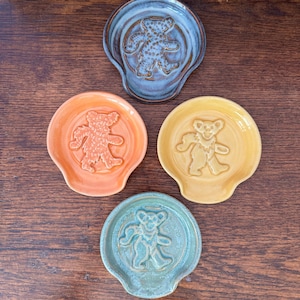 Puede incluir: Cuatro reposacucharas de cerámica con un diseño de oso. Los reposacucharas son en tonos de azul, naranja, amarillo y verde.