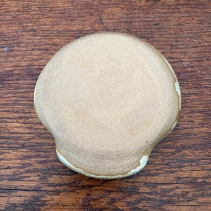 May include: A handmade, ceramic soap dish with a light tan color and a unique, organic shape. The dish has a slightly raised edge with a subtle, off-white glaze. The dish is designed to hold a bar of soap.