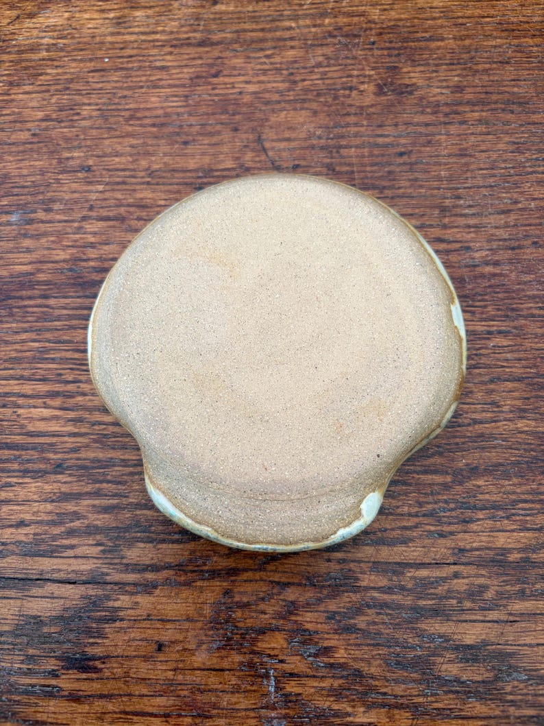 May include: A handmade, ceramic soap dish with a light tan color and a unique, organic shape. The dish has a slightly raised edge with a subtle, off-white glaze. The dish is designed to hold a bar of soap.