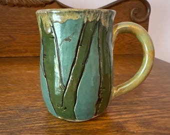 Könnte beinhalten: Eine handgefertigte Keramik-Tasse mit einem teal- und grünen Farbschema. Die Tasse hat ein strukturiertes Design aus vertikalen Linien und einen abgerundeten Henkel. Der Rand und der Henkel haben eine gelbgrüne Glasur.