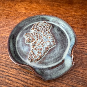 Puede incluir: Un reposacucharas de cerámica con un esmalte azul y verde. El reposacucharas tiene la forma de la isla de Irlanda y tiene un diseño de color dorado.