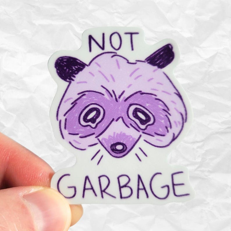 以下が含まれることがあります： 紫と白のステッカーに、漫画の浣腸の顔と「NOT GARBAGE」の文字が書かれています。